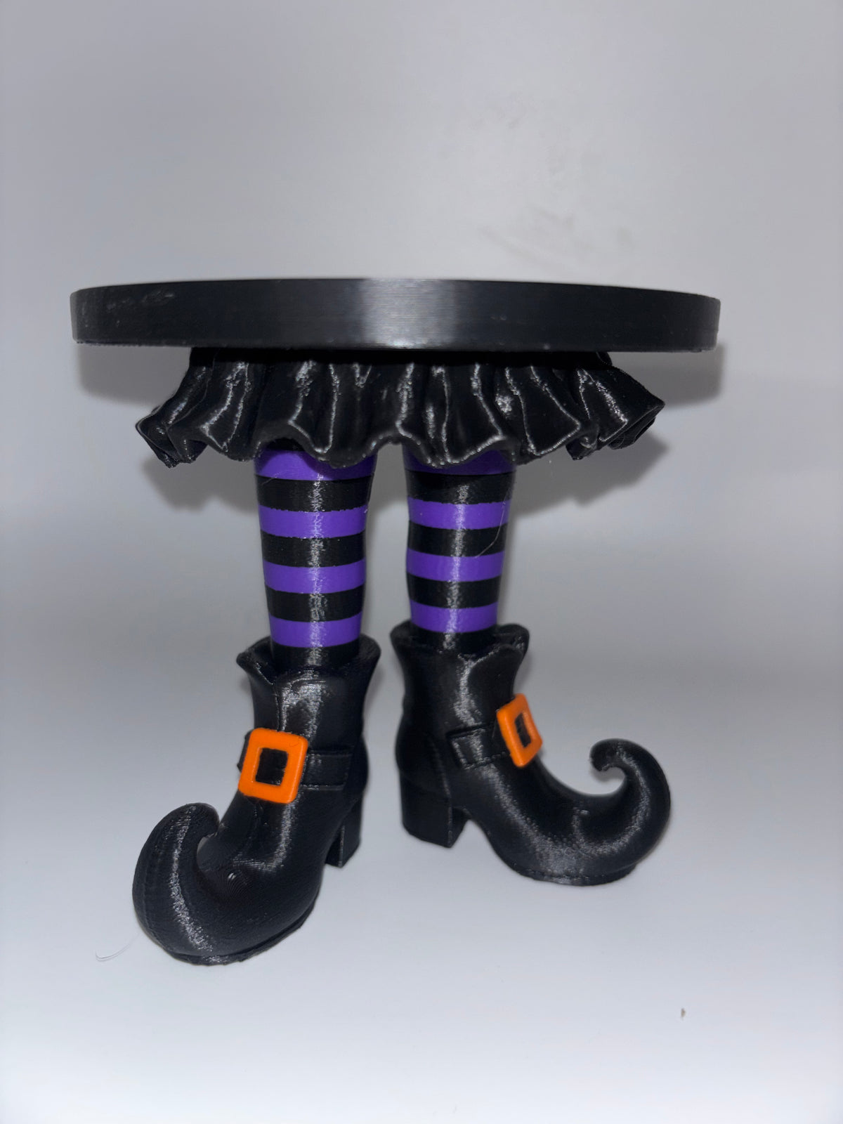 A Halloween-themed decorative figurine of two witch legs with black and purple striped stockings, black pointed boots with orange buckle details, and a black ruffled base, shown on a plain light gray background.